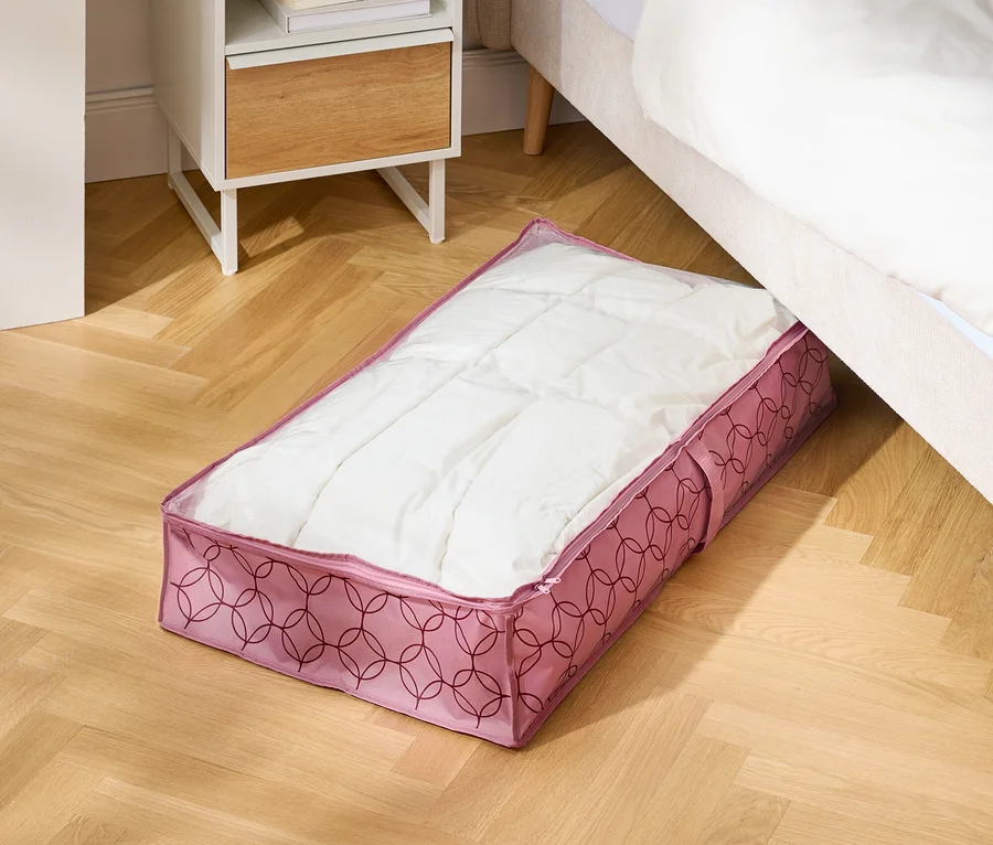 Sur un plancher en bois, un sac de rangement rose est placé sous un lit à côté d'une table de chevet.