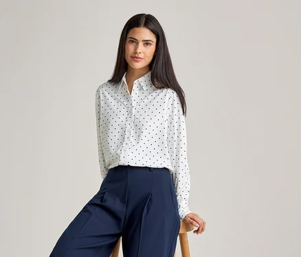 Une femme aux cheveux foncés porte un chemisier blanc à pois noirs et un pantalon bleu, assise sur un tabouret.