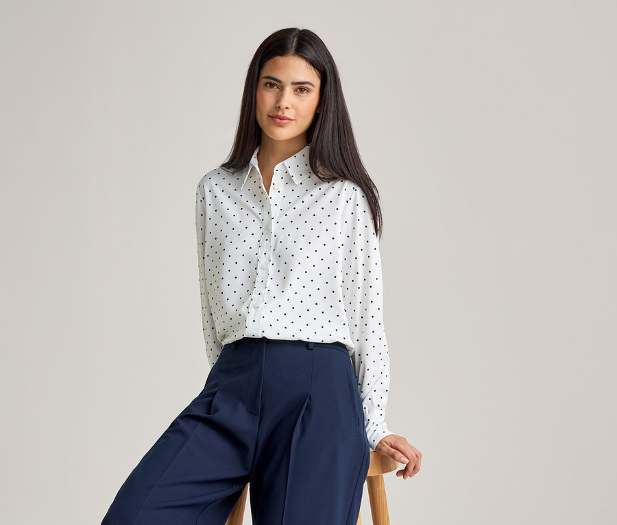 Une femme aux cheveux foncés porte un chemisier blanc à pois noirs et un pantalon bleu, assise sur un tabouret.