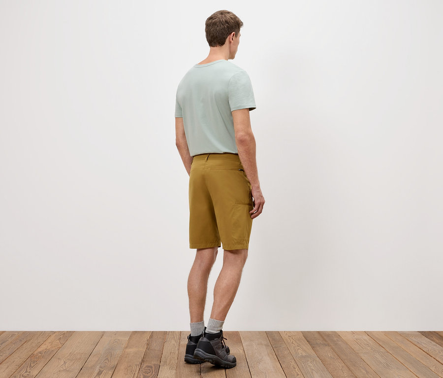 Un homme se tient dos à la caméra, portant un t-shirt vert clair, un short doré et des chaussures sombres sur un plancher en bois.