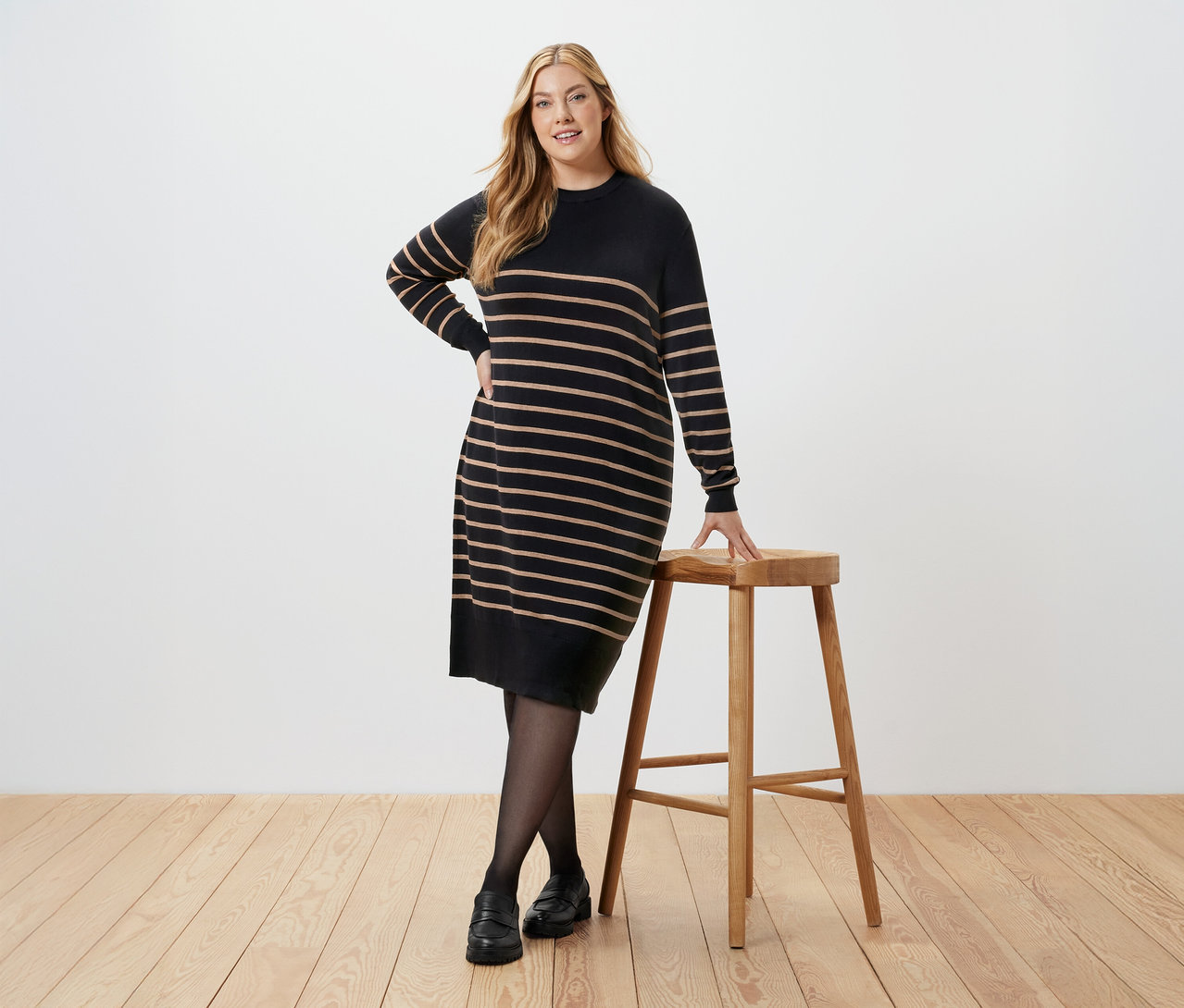 Femme posant en robe pull rayée à côté d'un tabouret.