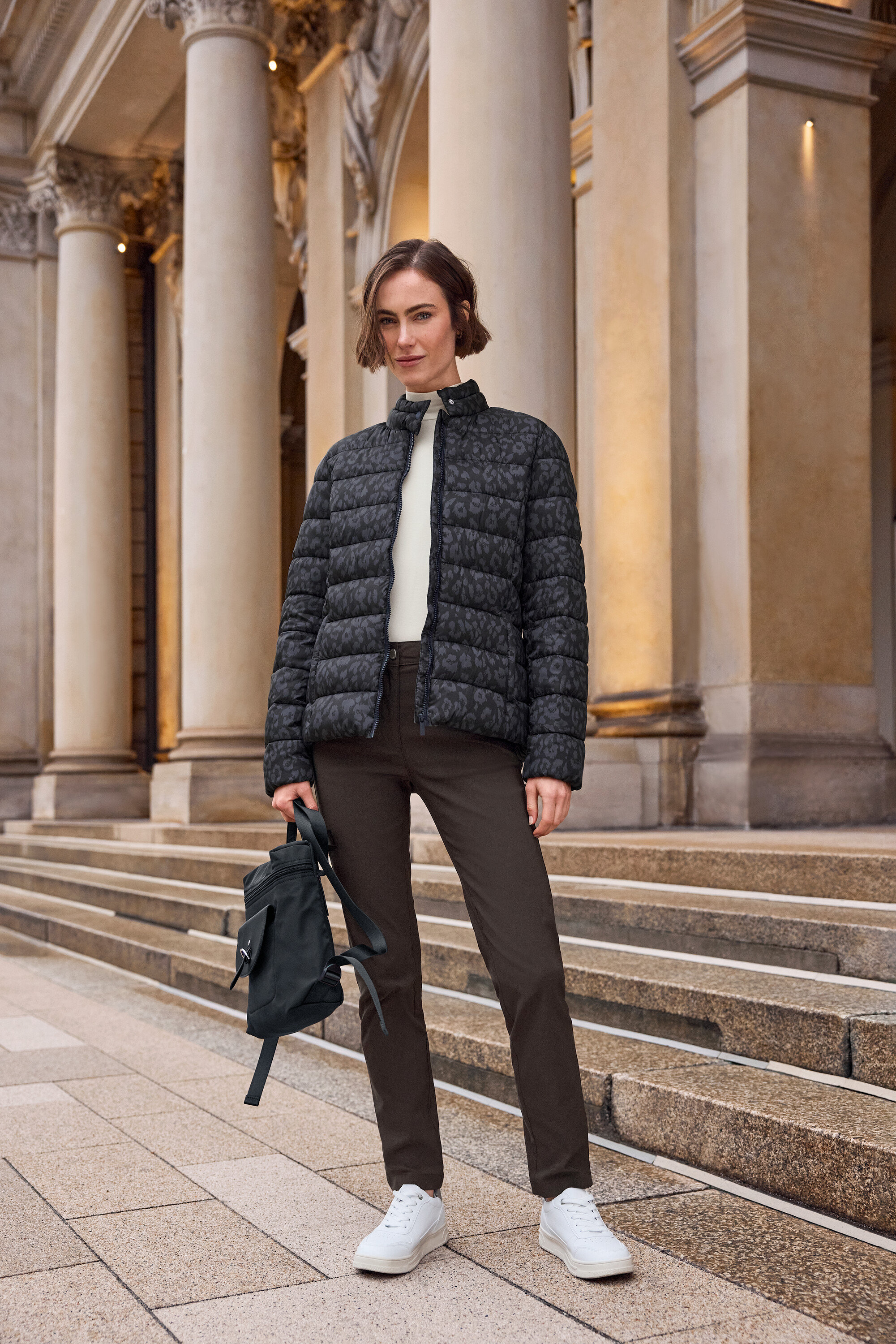 Femme debout devant un escalier, portant un blouson en duvet à motif léopard, un pantalon marron et des baskets blanches Tom Tailor en PU.