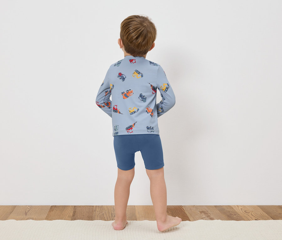 Un enfant, vu de dos, porte un t-shirt bleu à manches longues avec des dessins colorés de véhicules de construction et un short bleu.