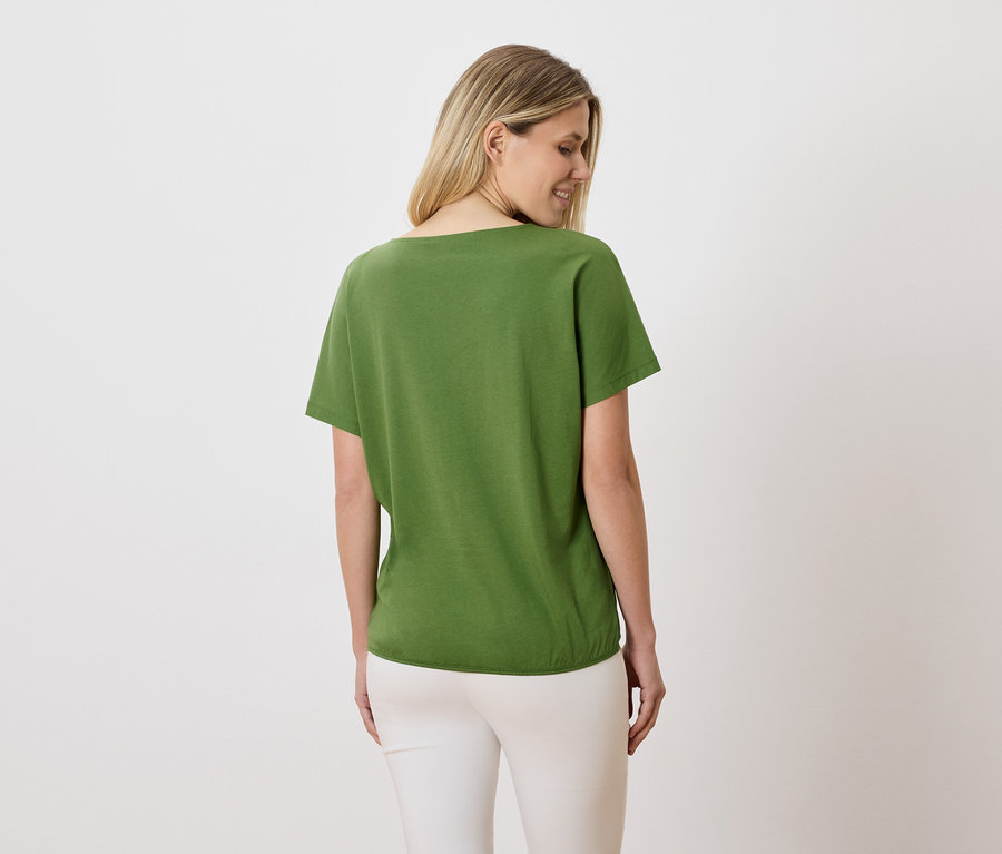 Une femme aux cheveux blonds porte un t-shirt vert et un pantalon blanc.
