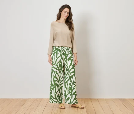 Femme debout devant un mur portant un haut beige et un pantalon vert ample à motifs de feuilles.