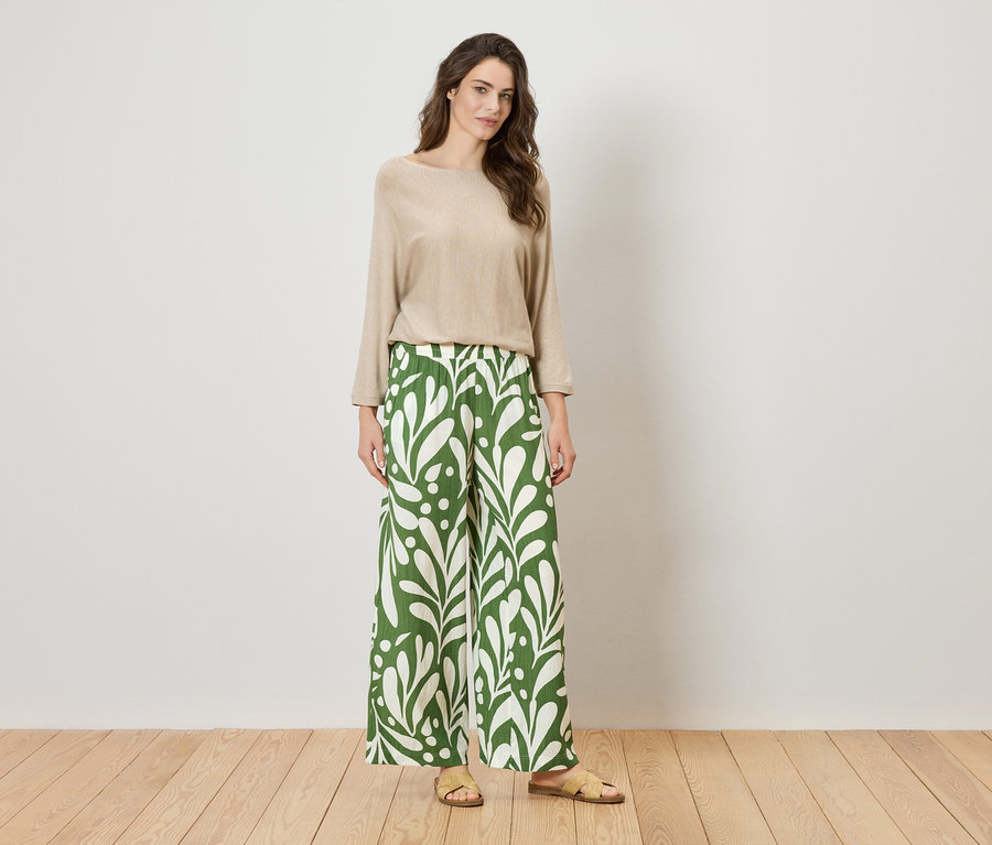 Femme debout devant un mur portant un haut beige et un pantalon vert ample à motifs de feuilles.