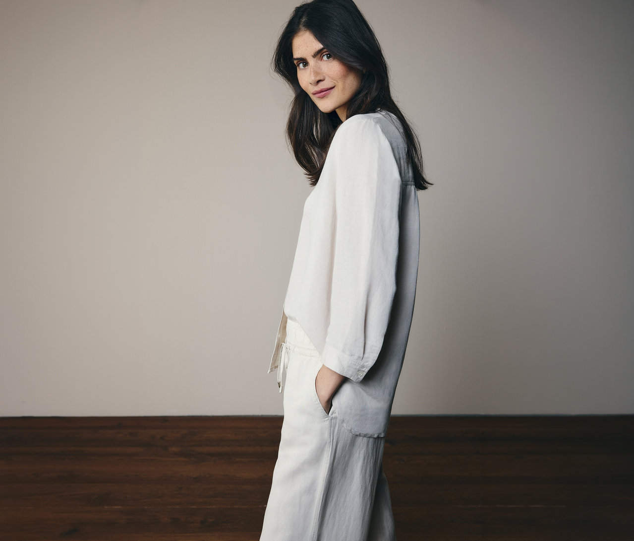 Une femme aux cheveux foncés pose en haut en lin blanc et pantalon assorti sur un fond clair.