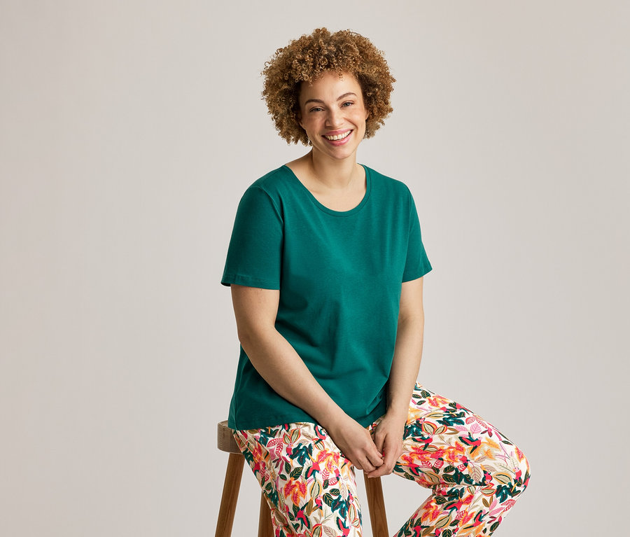 Femme souriante aux cheveux bouclés assise sur un tabouret en bois, portant un t-shirt vert et un pantalon de pyjama fleuri d'un ensemble de pyjama.