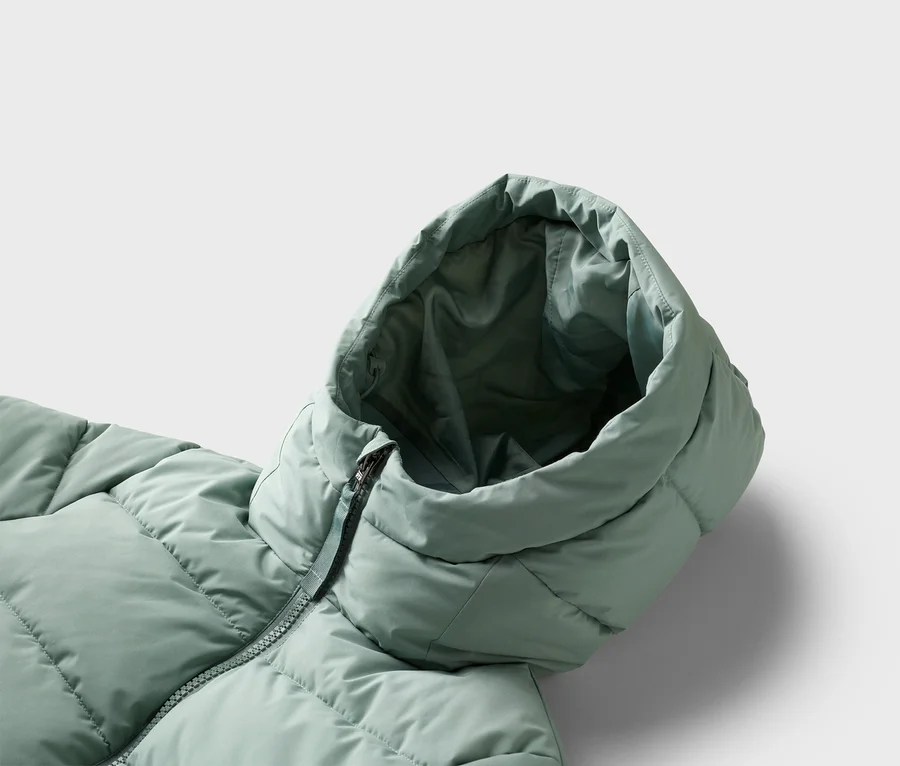 Gros plan d'un manteau matelassé vert clair avec capuche et fermeture éclair sur un fond gris clair.