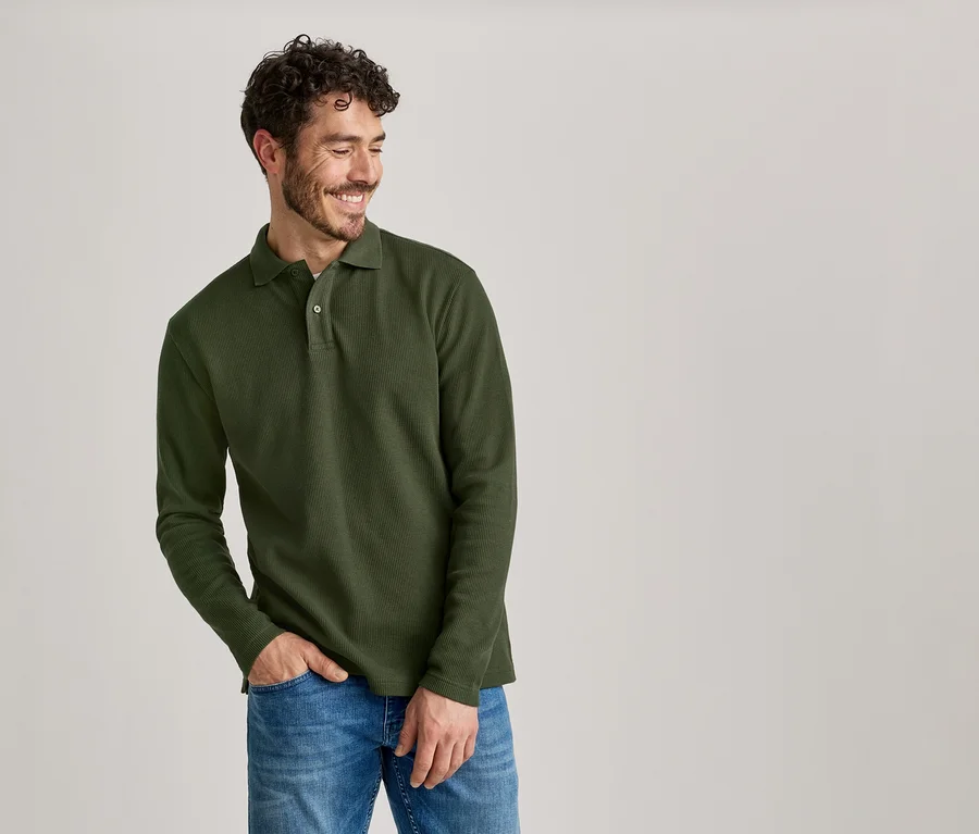 Homme souriant aux cheveux bouclés portant un polo olive à manches longues et un jean, une main dans la poche.