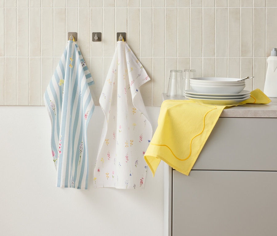 Deux torchons suspendus à des crochets sur un mur au-dessus d'un comptoir de cuisine, sur lequel se trouvent de la vaisselle, des verres, un flacon de liquide vaisselle et un chiffon jaune.