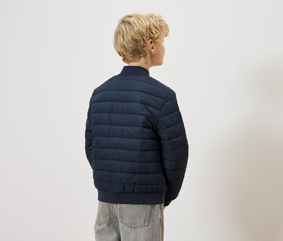 Vue arrière d'un enfant blond portant un blouson matelassé bleu marine et un jean gris sur un fond blanc.