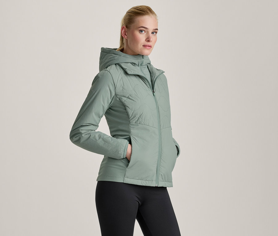 Femme portant une veste à capuche vert clair et un legging noir, les mains dans les poches de la veste.