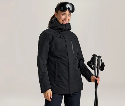 Femme souriante portant une veste de ski noire et des lunettes de ski sur la tête, tenant des bâtons de ski noirs.