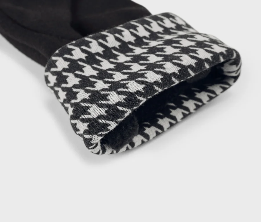 Gros plan d'une manchette de vêtement noir, repliée pour révéler un motif pied-de-poule noir et blanc. La doublure intérieure est sombre et douce.