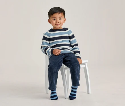 Jeune garçon assis sur un tabouret blanc, portant un pull rayé bleu, bleu clair et blanc, un pantalon en velours côtelé bleu foncé et des chaussettes rayées, souriant à la caméra.