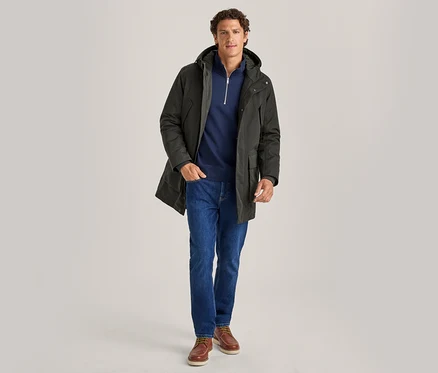 Homme debout, portant une parka à capuche vert foncé, un pull zippé bleu marine, un jean bleu et des chaussures bateau marron, les mains dans les poches.