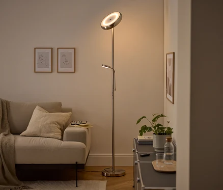 Un lampadaire argenté moderne avec une lumière principale orientée vers le haut et une liseuse réglable éclaire un salon confortable. Un canapé beige avec un plaid et un coussin, deux tableaux d'art linéaire encadrés et un meuble sombre avec une plante et une bouteille d'eau sont visibles.
