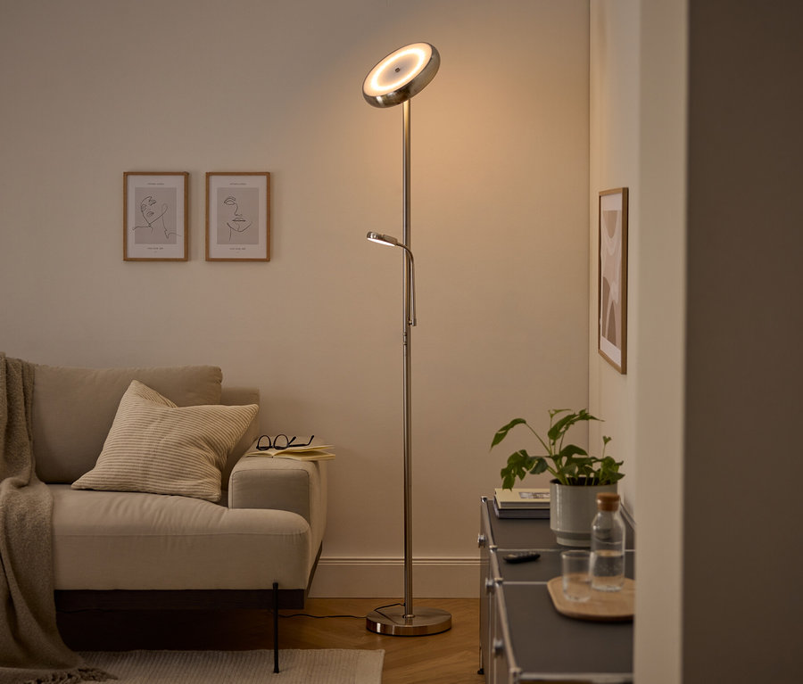 Un lampadaire argenté moderne avec une lumière principale orientée vers le haut et une liseuse réglable éclaire un salon confortable. Un canapé beige avec un plaid et un coussin, deux tableaux d'art linéaire encadrés et un meuble sombre avec une plante et une bouteille d'eau sont visibles.