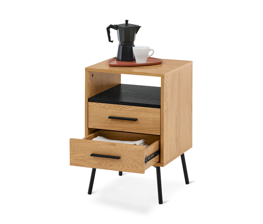 Table de chevet en bois clair avec des accents noirs et des pieds inclinés. Sur le dessus, une cafetière moka noire et une tasse blanche sont posées sur un plateau brun-rouge. En dessous, une étagère ouverte avec un intérieur noir est suivie de deux tiroirs, le tiroir inférieur étant ouvert et révélant du linge blanc plié.
