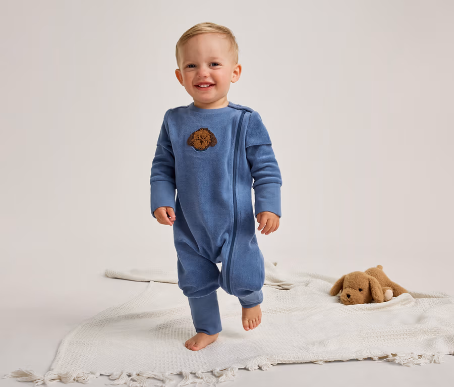 Un bébé souriant se tient pieds nus sur une couverture blanche. Il porte une grenouillère bleue à manches longues avec une tête de chien marron brodée. À droite, un chien en peluche marron est posé sur la couverture.