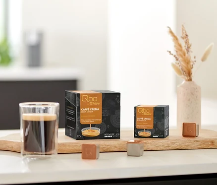 Un verre de café avec de la crème, deux paquets de capsules Qbo Caffè BUNA ENTETA (8 et 27 capsules) et deux capsules individuelles sur une planche en bois.