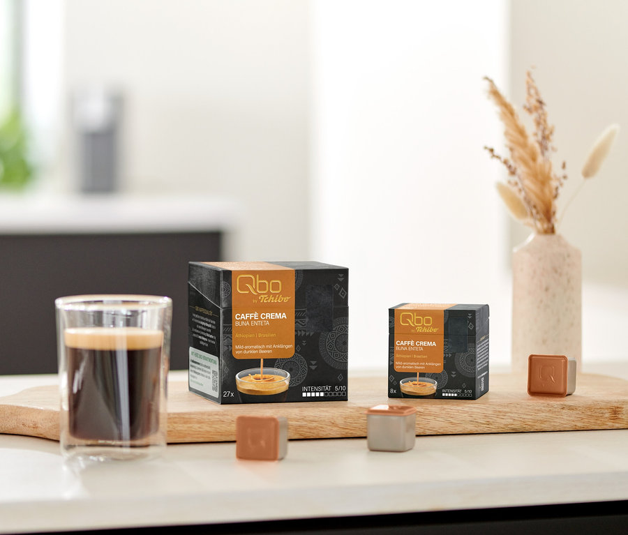 Un verre de café, une boîte de 27 et une boîte de 8 capsules Qbo Caffè BUNA ENTETA, ainsi que deux capsules Qbo individuelles sont posés sur une planche en bois. Un bol de biscuits et un vase de fleurs séchées sont en arrière-plan.