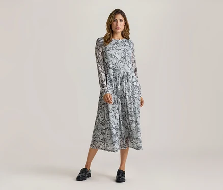 Femme debout, portant une robe midi à manches longues avec un motif peau de serpent gris et noir et des mocassins noirs.