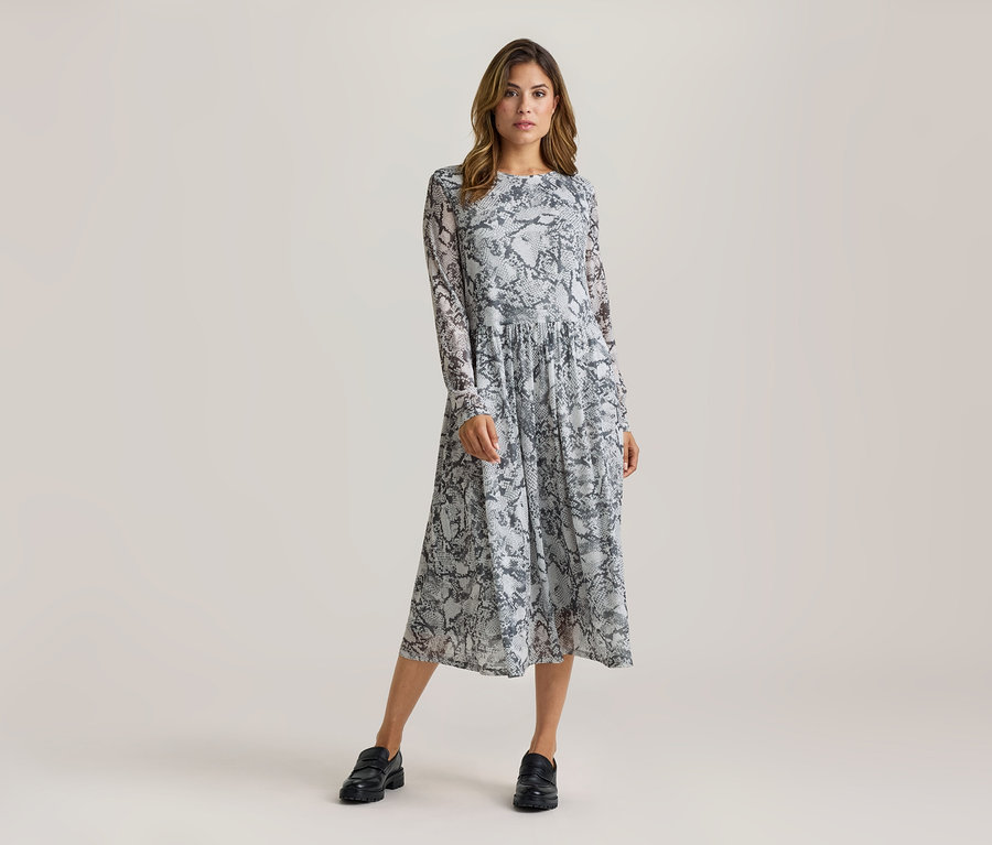 Femme debout, portant une robe midi à manches longues avec un motif peau de serpent gris et noir et des mocassins noirs.