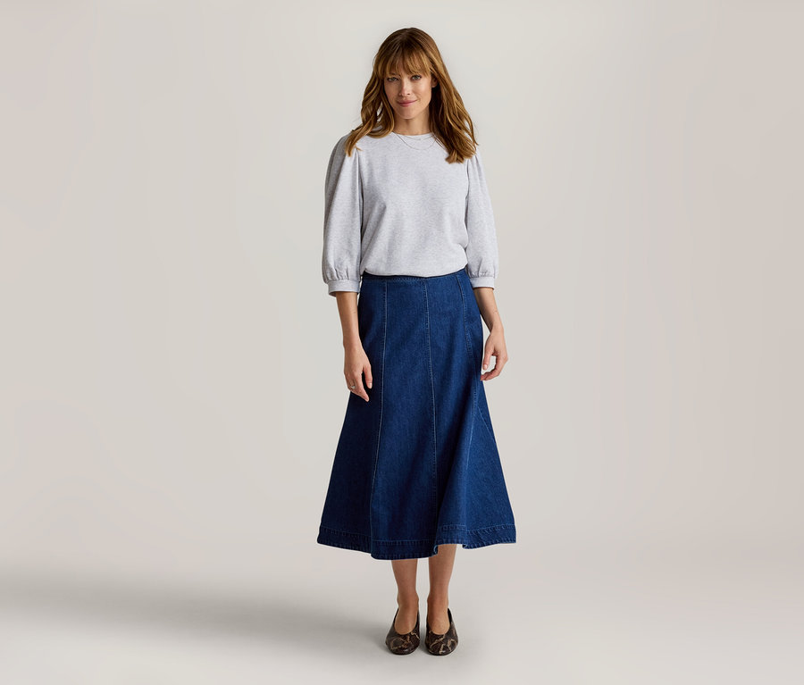 Femme debout, portant un haut gris clair à manches bouffantes trois-quarts, une longue jupe en jean bleu foncé et des chaussures plates foncées à motifs.