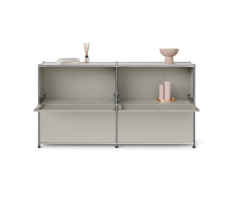 Meuble modulaire gris clair avec cadre chromé, comprenant deux compartiments supérieurs ouverts et deux portes inférieures abattantes ouvertes. Les étagères supérieures présentent des livres, un diffuseur de parfum, un bol en bois et deux bougies roses sur un plateau doré.