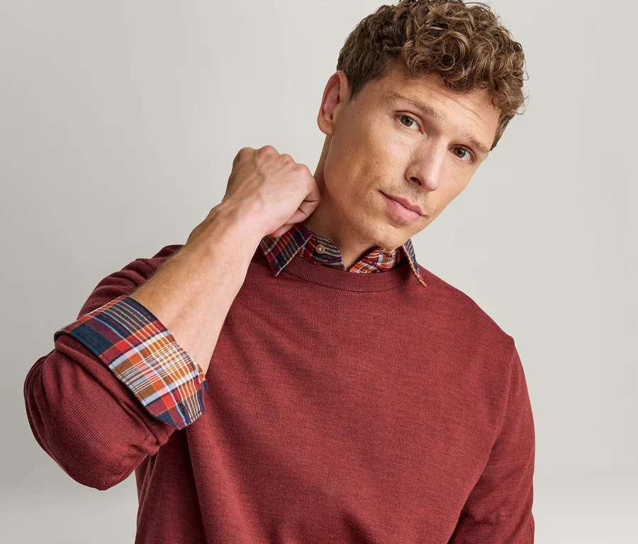 Homme portant un pull rouge sur une chemise à carreaux, avec le col et les poignets retroussés de la chemise visibles. Il regarde l'appareil photo.