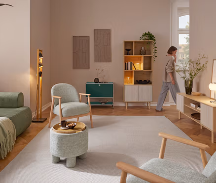 Une femme traverse un salon meublé d'un canapé vert, de deux fauteuils avec structure en bois et d'un pouf avec tablette. Un lampadaire Trio « Bell » se tient à côté du canapé. Contre le mur du fond se trouvent un buffet en métal « CN3 » et une bibliothèque aspect travertin avec deux lampes de table LED Trio « Madison ». À droite, un meuble bas aspect travertin avec une lampe de table LED Trio « Madison » et un vase de fleurs.