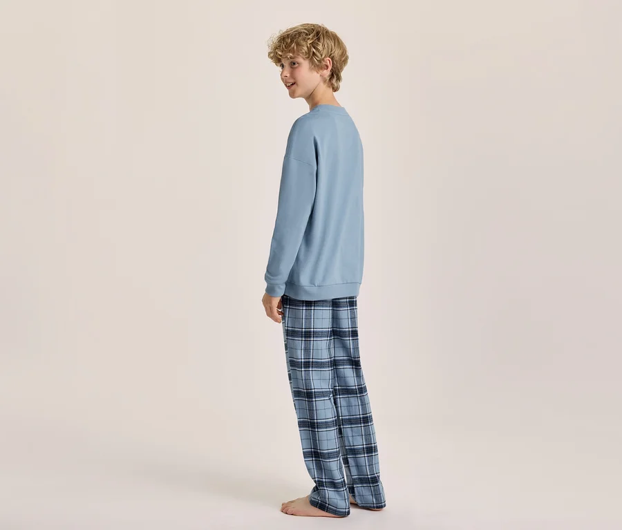 Un jeune garçon aux cheveux blonds bouclés, portant un sweat-shirt bleu clair à manches longues et un pantalon de pyjama à carreaux bleus, se tient pieds nus, regardant par-dessus son épaule gauche.