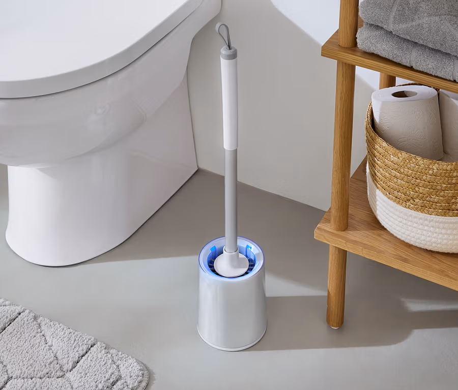 Dans la salle de bain, des toilettes blanches sont à côté d'une brosse WC avec une lumière bleue et d'une étagère en bois avec des serviettes et du papier toilette.