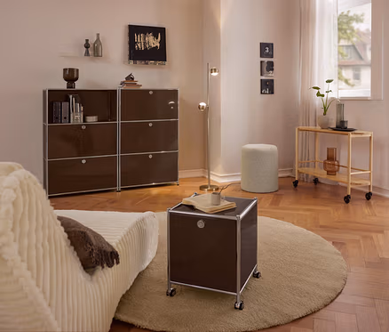 Salon moderne avec canapé beige, grande commode métallique marron »CN3« à 3 compartiments rabattables, petite commode métallique roulante marron »CN3« à 2 compartiments rabattables sur tapis rond, pouf bouclé et chariot de service en bois.