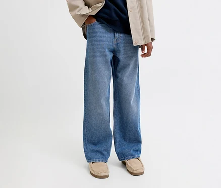 Personne portant un jean large bleu, une veste beige, un haut bleu marine et des mocassins beiges. Une main est dans la poche.