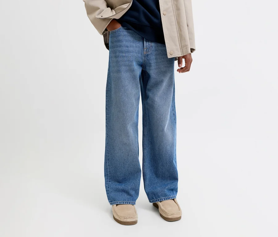 Personne portant un jean large bleu, une veste beige, un haut bleu marine et des mocassins beiges. Une main est dans la poche.