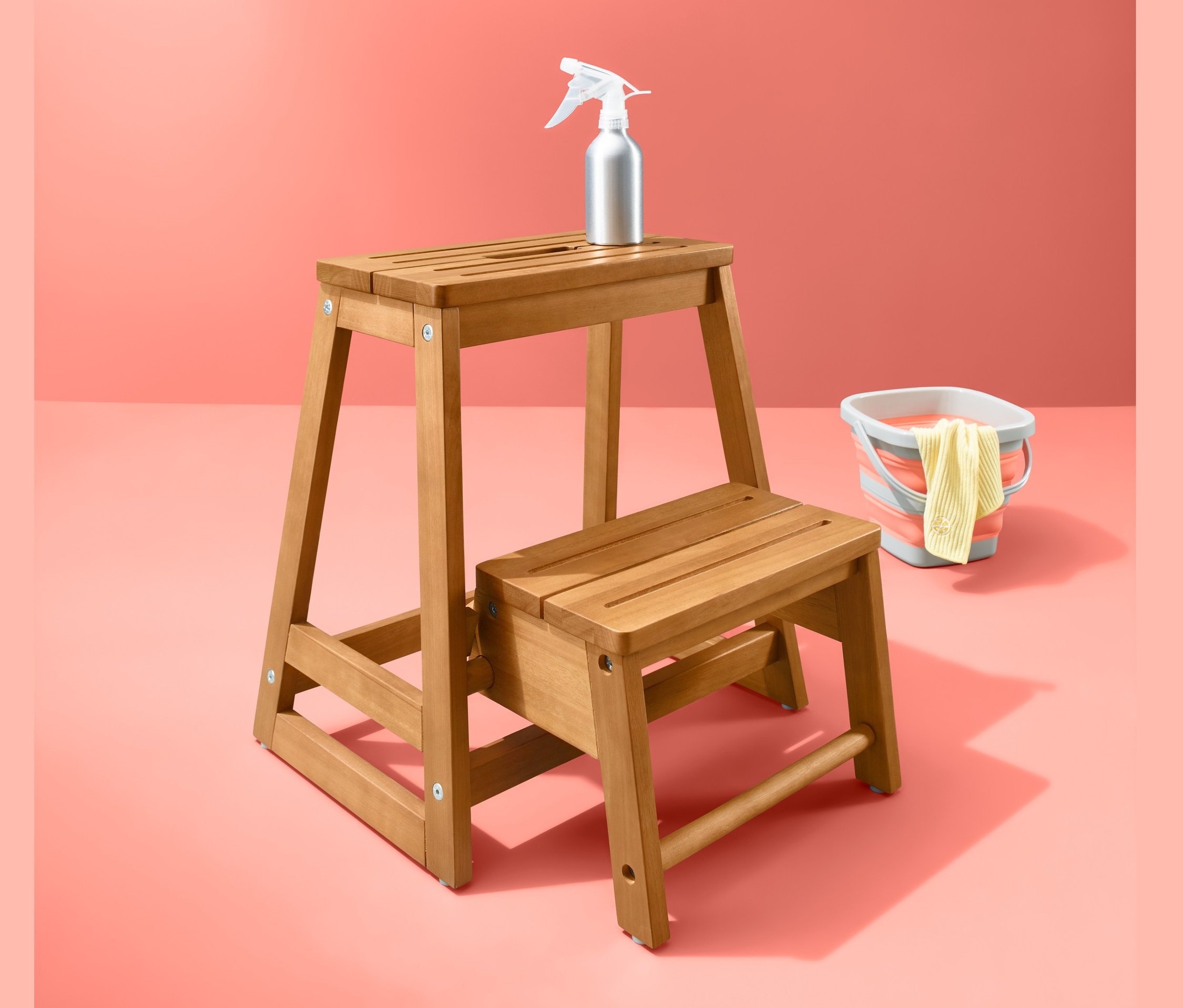 Un marchepied pliant en bois avec un vaporisateur et un seau sur un fond corail.