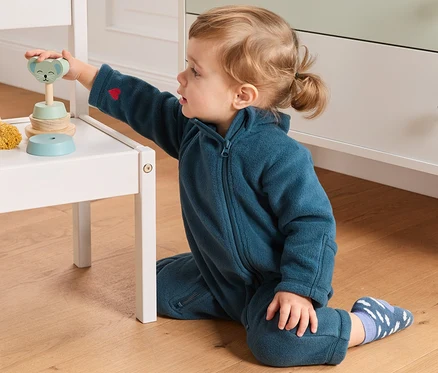 Un bébé joue avec un jouet en bois sur une chaise blanche, il porte une combinaison en polaire en matériaux recyclés.