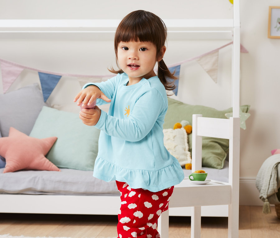 Une fille se tient dans une chambre d'enfant et porte un t-shirt bleu clair et un pantalon rouge avec des nuages blancs. Elle porte 3 t-shirts à manches longues en coton bio.