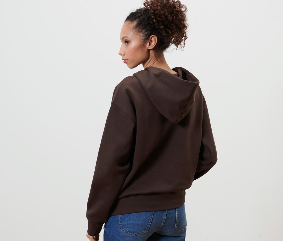 Une femme pose dos à la caméra et porte un sweat-shirt à capuche marron foncé.