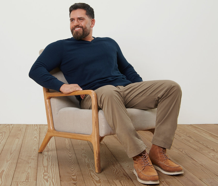 Un homme est assis dans un fauteuil et porte un pantalon chino sable et un pull bleu marine à col en V.