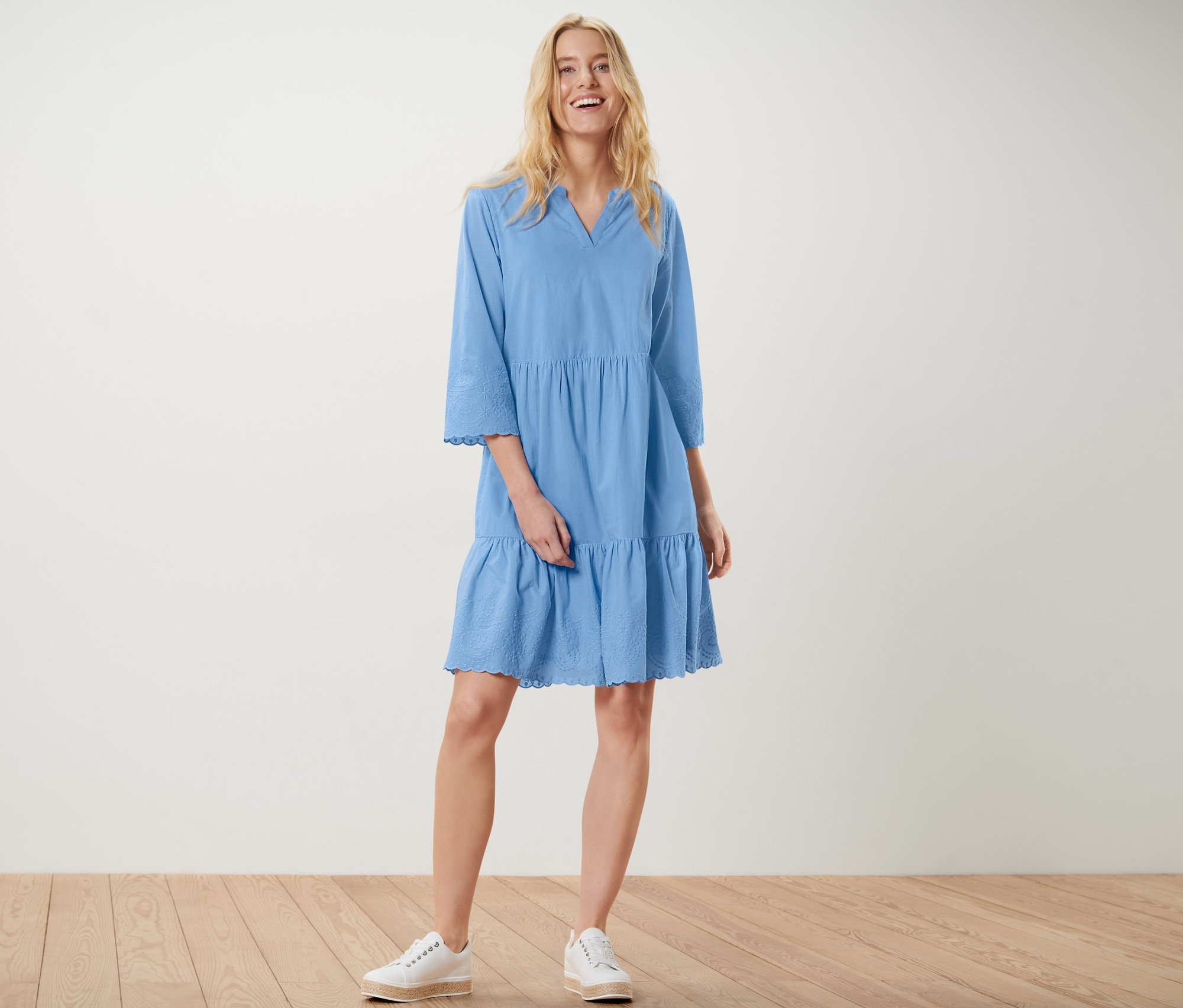 Une femme blonde sourit et porte une robe tissée bleue avec des broderies et des baskets blanches.