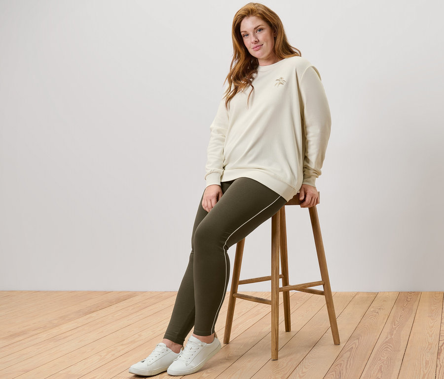 Femme assise sur un tabouret portant un sweatshirt avec broderie et un legging passepoilé.