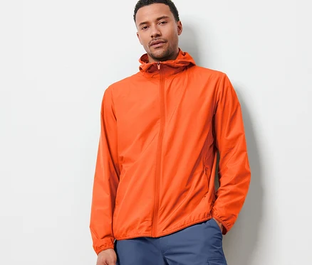 Un homme portant une veste technique légère « packable » orange et un pantalon bleu pose devant un mur blanc.