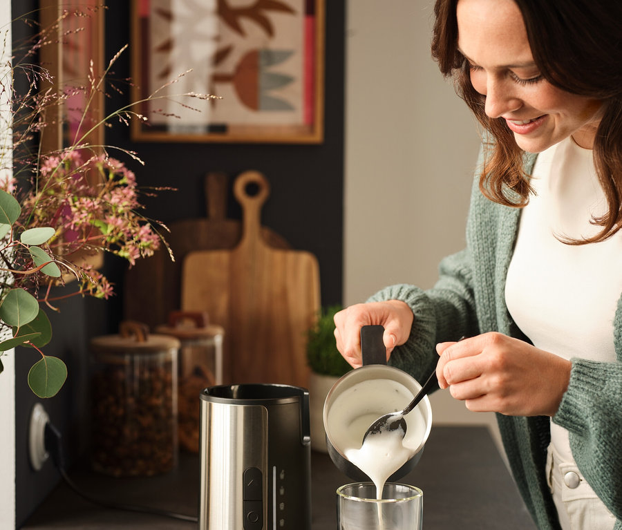 Une femme verse de la mousse de lait avec une cuillère dans un verre de café à côté d'un mousseur à lait.