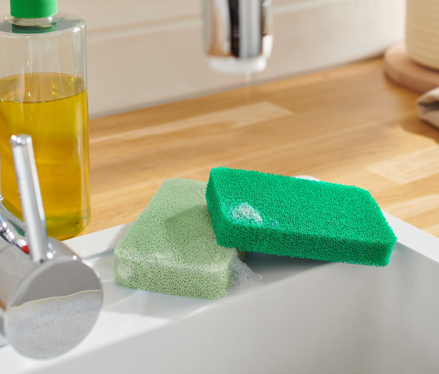 Deux éponges en silicone sont posées dans un évier blanc à côté d'un robinet et d'un flacon de produit de nettoyage.