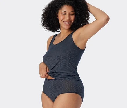 Femme souriante aux cheveux bouclés portant un débardeur SCHIESSER bleu foncé à rayures et un slip midi assorti avec modal. Elle remonte son débardeur d'une main.