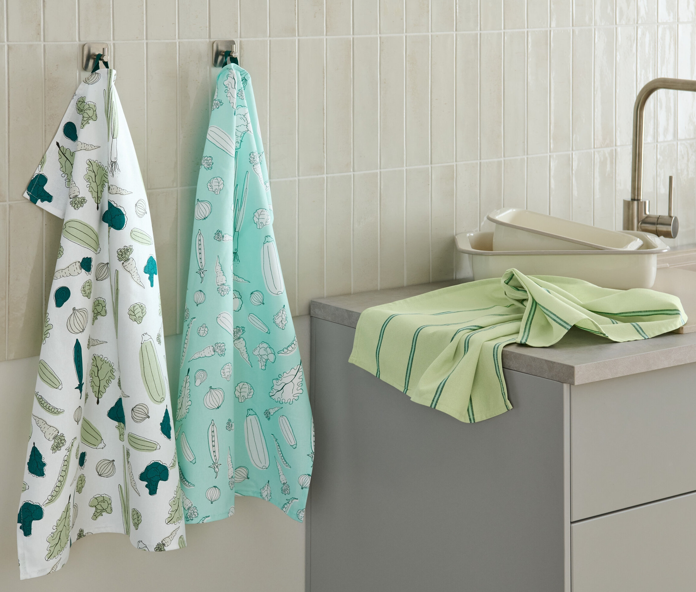 Deux linges de cuisine à motifs de légumes sont accrochés au mur, un autre linge de cuisine vert est posé sur le plan de travail.